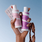 A hand holds two Boucléme haircare bottles against a light blue gradient background, with a curl cream bottle on the left in pink and a super hold styler bottle on the right in purple. Tortoiseshell sunglasses hang from the hand, and printed cards are tucked behind the bottles.