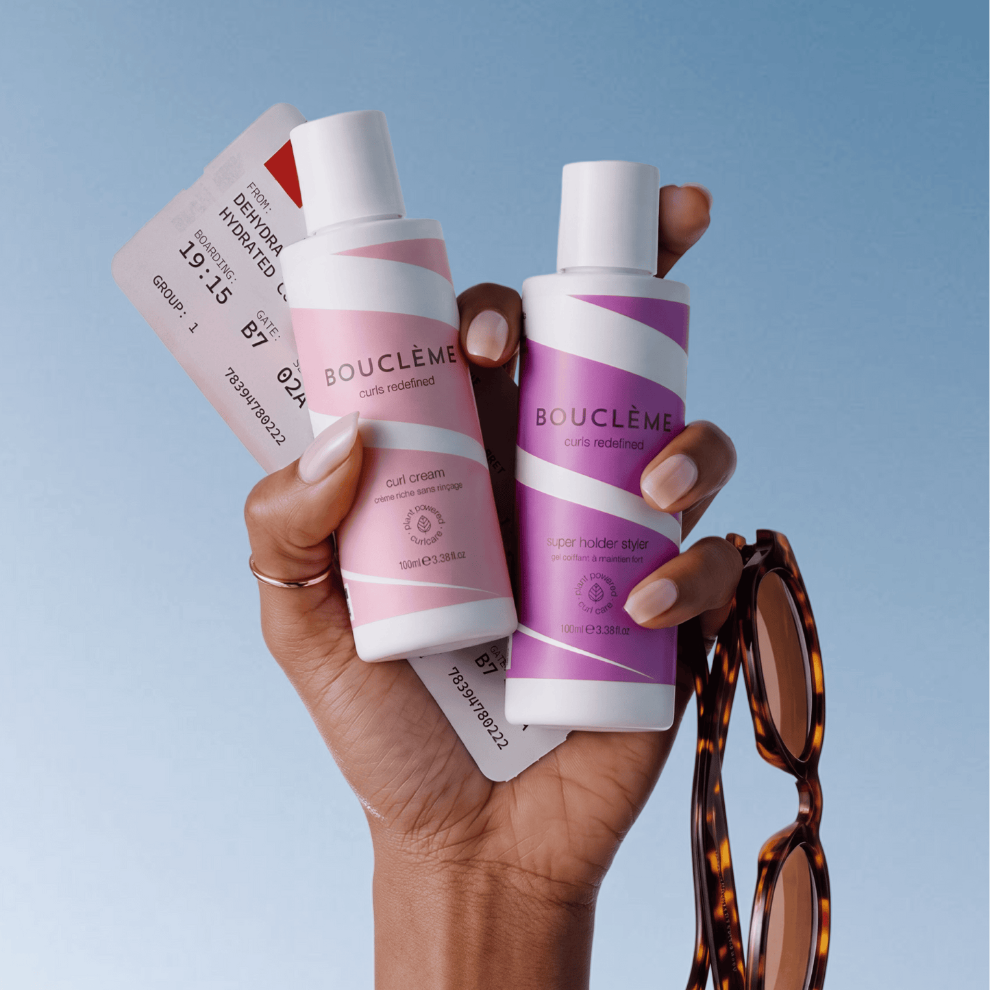 A hand holds two Boucléme haircare bottles against a light blue gradient background, with a curl cream bottle on the left in pink and a super hold styler bottle on the right in purple. Tortoiseshell sunglasses hang from the hand, and printed cards are tucked behind the bottles.