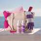 Bouclemi Hydration & Hold Duo displayed in front of a beige tote bag with beach accessories, including a pink bikini top, striped towel, and purple-and-white fringed towel, set against an ocean background.